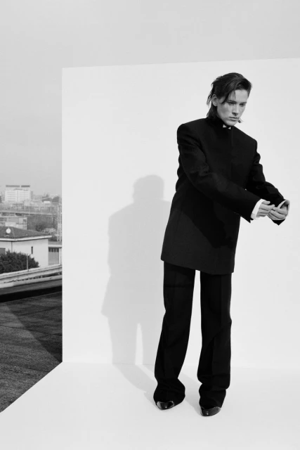 Black and white studio shot: Person in oversized black suit poses in crisp white corner setting with a city building background.