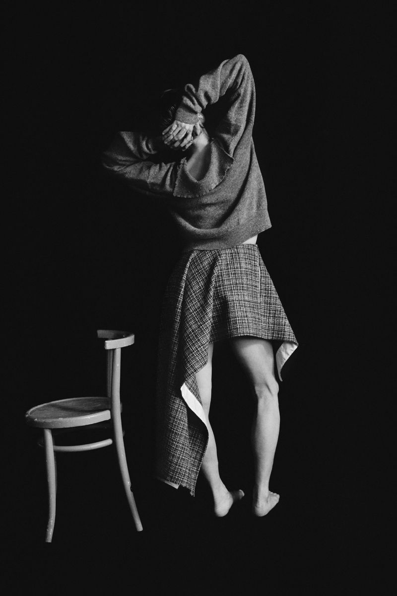 Model standing with back to camera wearing a grey sweater and patterned skirt next to a chair on black