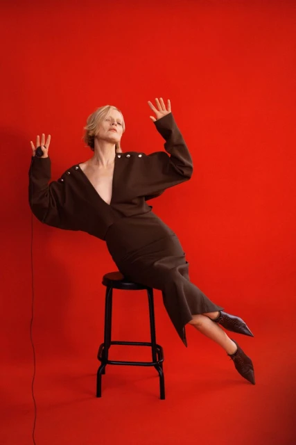 Blonde woman in a brown dress posing on a stool against a red backdrop, her hands raised, creating a high-fashion studio portrait.