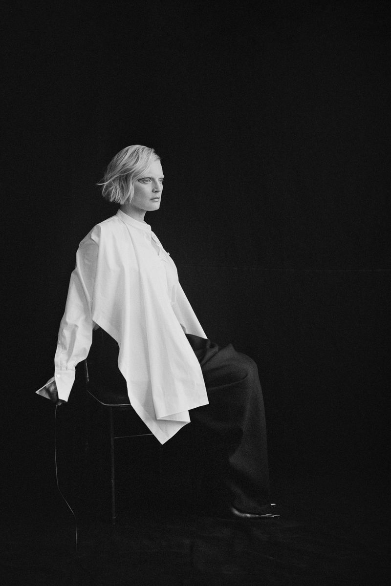 Black and white portrait of a woman with short hair seated in profile on a chair wearing an asymmetrical draped white shirt and dark trousers.