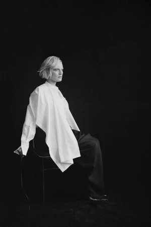 Black and white portrait of a woman with short hair seated in profile on a chair wearing an asymmetrical draped white shirt and dark trousers.