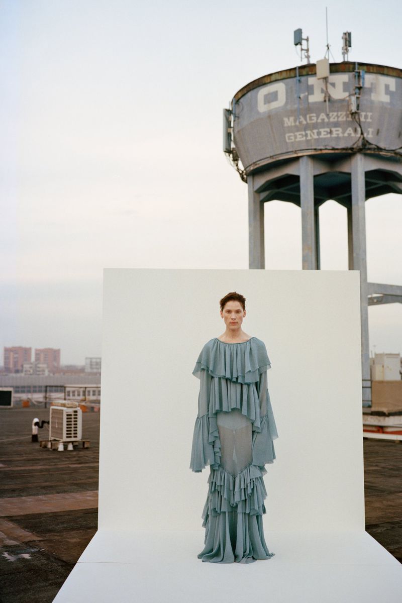 Fashion photography featuring a woman in a light blue ruffled dress standing on a white platform against an industrial cityscape backdrop.