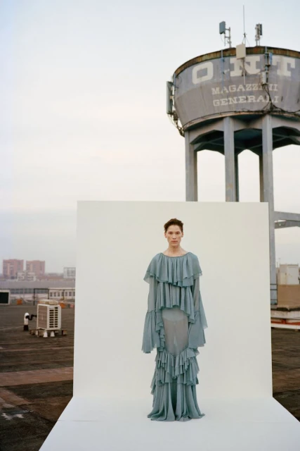 Fashion photography featuring a woman in a light blue ruffled dress standing on a white platform against an industrial cityscape backdrop.