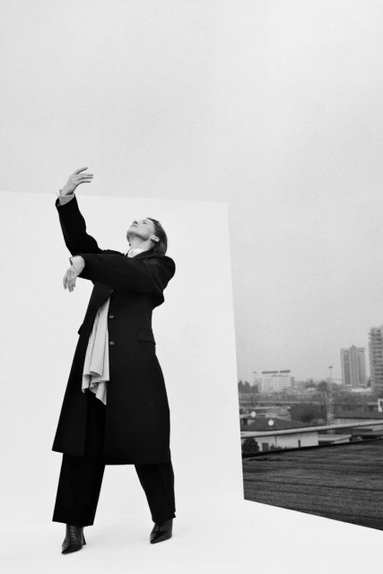 A person in a black coat and wide-leg pants stands next to a white wall with a cityscape in the background in a black and white editorial.