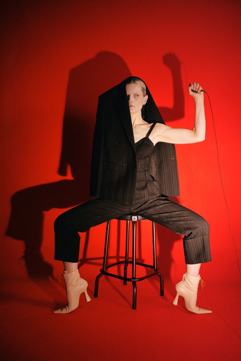 Fashion shot of an adult woman in a suit and high heels, seated on a stool against a red background. Studio lighting and unique styling.