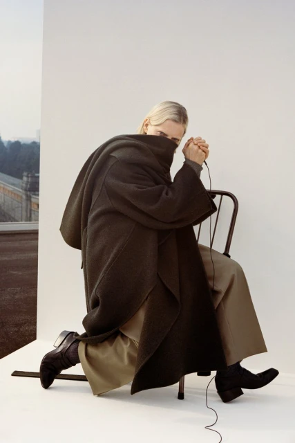 Fashion shot of a blond woman in a dark coat kneeling on a chair, with an outdoor landscape visible in the background.