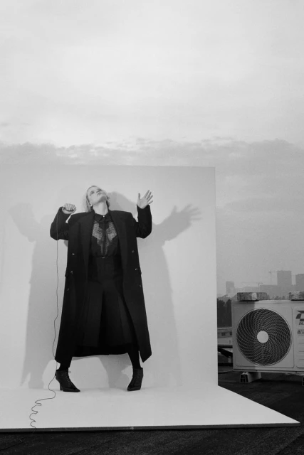 Monochrome fashion shot of a woman in a coat, standing on a white platform against a cityscape backdrop. She's singing into a mic.