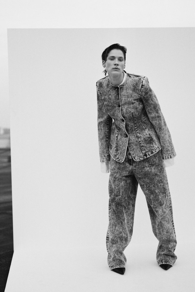 Monochrome fashion shot of a woman in a denim suit, posing against a backdrop of white and a dark landscape under an overcast sky.