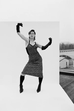 Model posing expressively in a textured midi dress, long black gloves, and pointed heels against an outdoor white backdrop.
