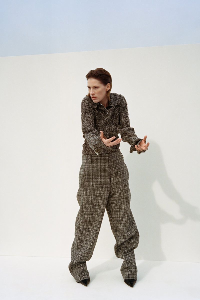 A woman in a brown tweed pantsuit poses against a split white and blue background in a fashion editorial-style studio shot.
