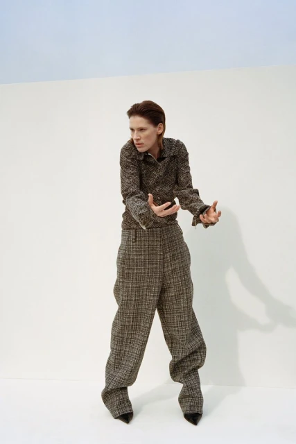 A woman in a brown tweed pantsuit poses against a split white and blue background in a fashion editorial-style studio shot.