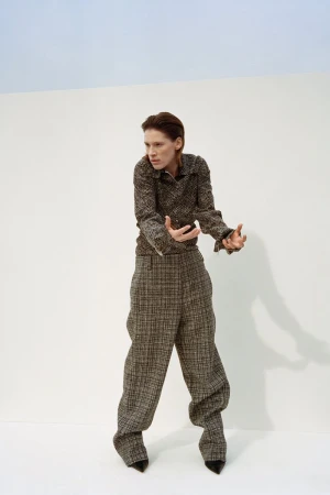 A woman in a brown tweed pantsuit poses against a split white and blue background in a fashion editorial-style studio shot.