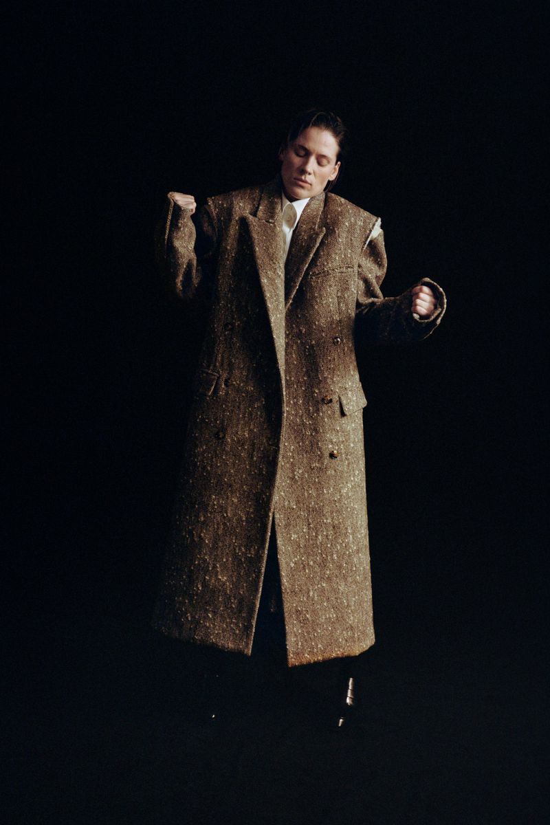 A person in a stylish, oversized brown tweed coat dances against a dark background in a studio setting, illuminated by directed light.