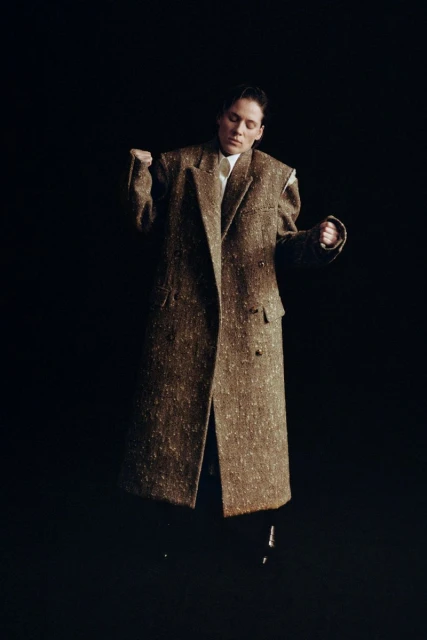 A person in a stylish, oversized brown tweed coat dances against a dark background in a studio setting, illuminated by directed light.