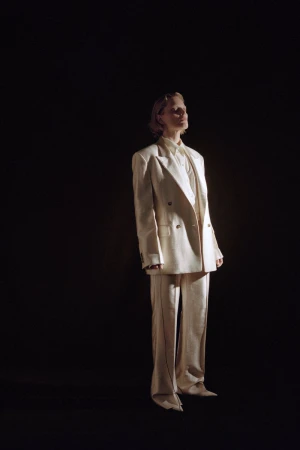 Model standing in an oversized cream double-breasted tailored suit against a solid black background under directional spotlighting.