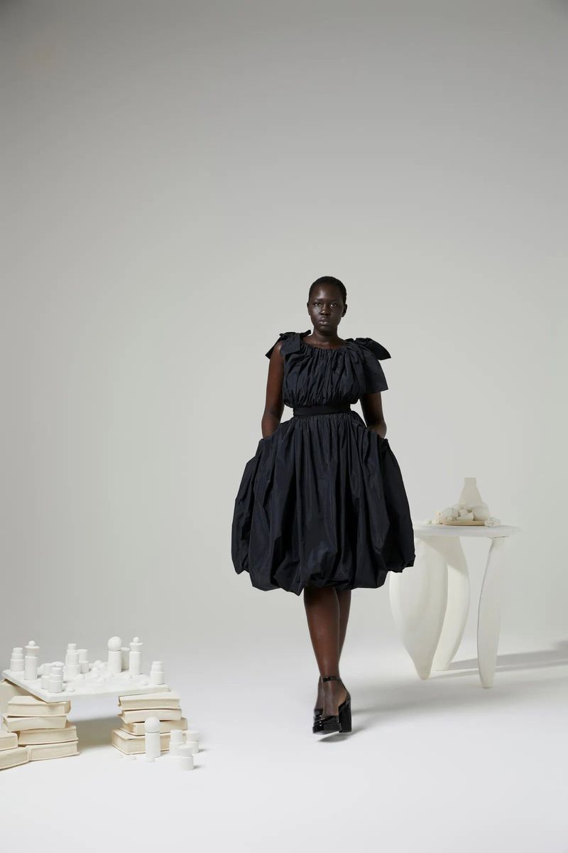 Full-length shot of a Black woman in a black dress and heels, striking a pose in a studio setting with artful objects.