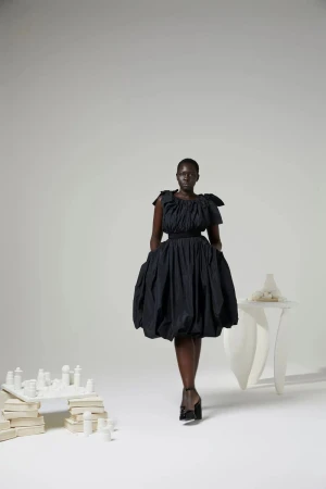 Full-length shot of a Black woman in a black dress and heels, striking a pose in a studio setting with artful objects. 