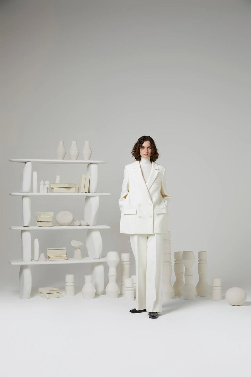 A woman with wavy brown hair stands in a white suit, complemented by a shelf filled with white vases in a bright studio setting.