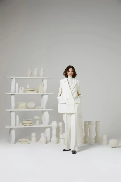A woman with wavy brown hair stands in a white suit, complemented by a shelf filled with white vases in a bright studio setting.