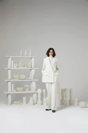 A woman with wavy brown hair stands in a white suit, complemented by a shelf filled with white vases in a bright studio setting.