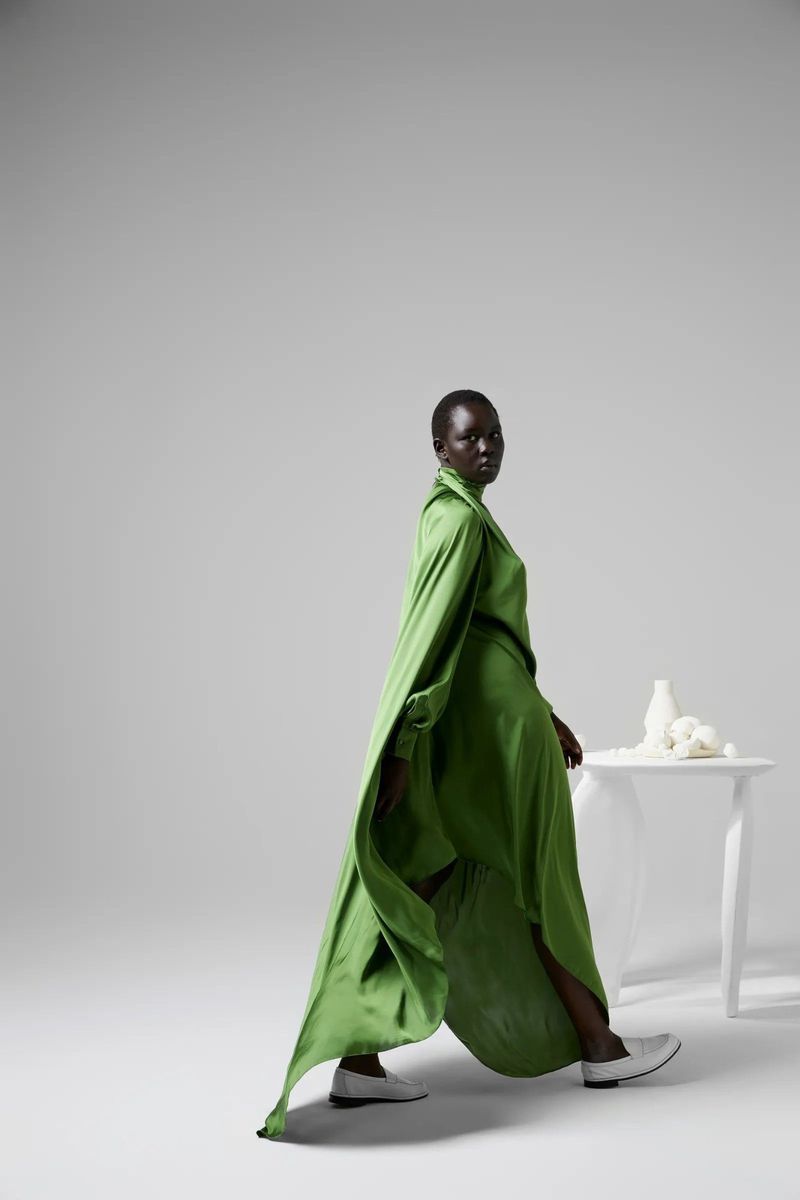 A model in a long green dress and white shoes walks toward the camera in a studio with a white table and vases in the background.