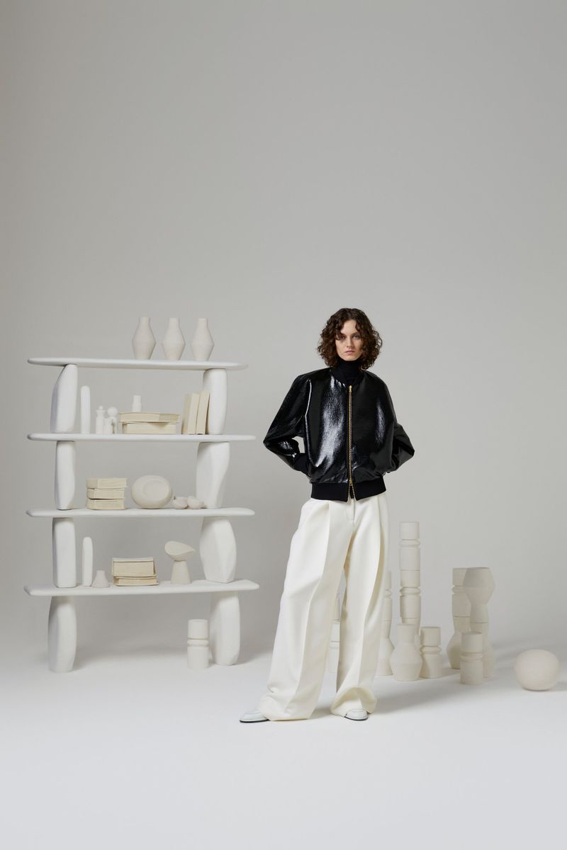 Fashion shot of a woman in a black jacket and white pants posing indoors, with a sculptural white bookshelf and decor behind her.