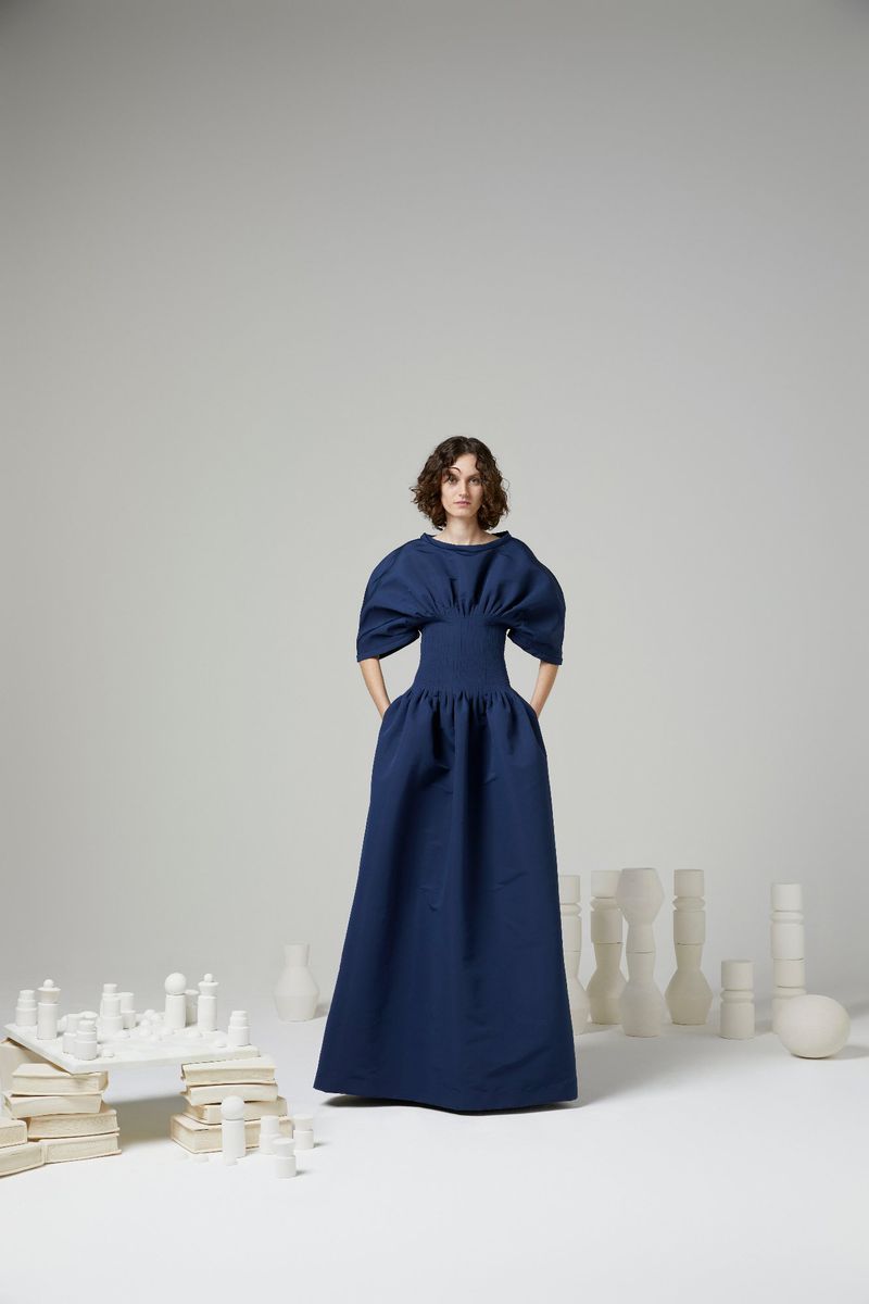 Adult female model stands in a full-length navy dress in a studio setting, surrounded by white, abstract chess-piece-like objects.