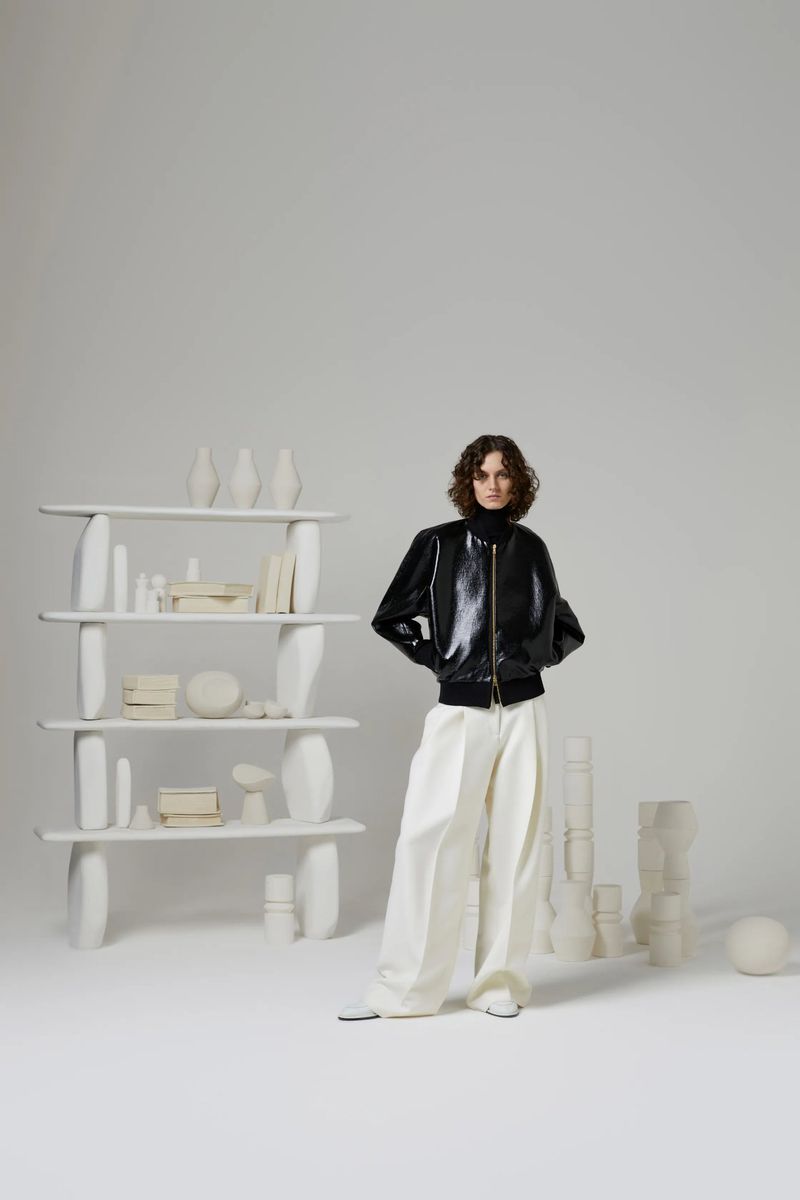 Indoor studio shot of a female fashion model wearing a black bomber jacket and loose white pants standing in front of a shelf and white objects.
