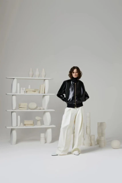 Indoor studio shot of a female fashion model wearing a black bomber jacket and loose white pants standing in front of a shelf and white objects.