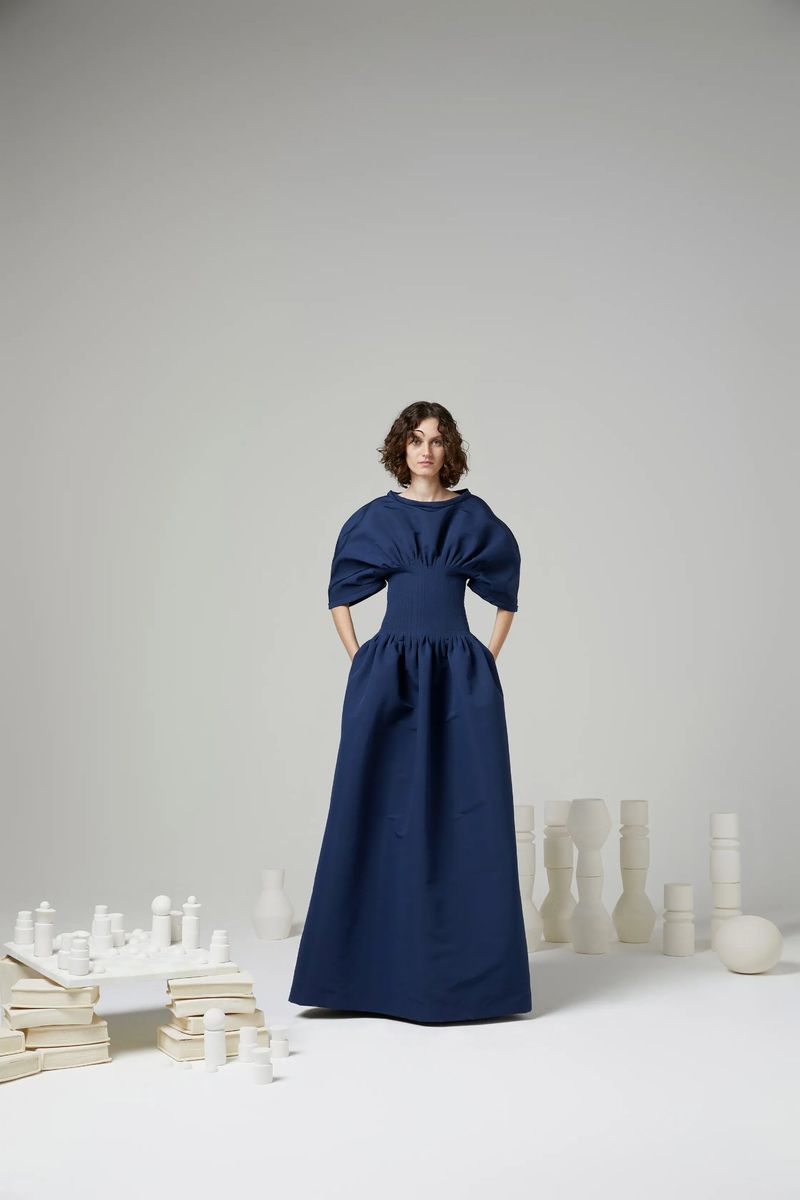Woman in a puffy-sleeved navy dress poses in a studio setting surrounded by white sculptural objects, creating a minimalist fashion image.