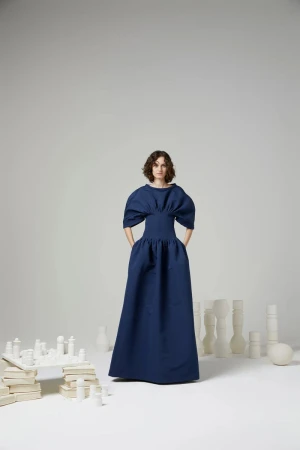 Woman in a puffy-sleeved navy dress poses in a studio setting surrounded by white sculptural objects, creating a minimalist fashion image.