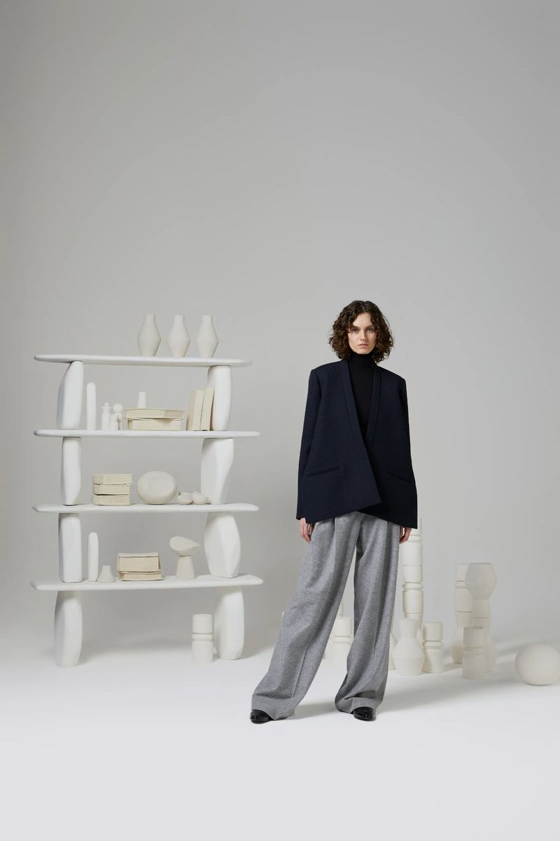 A woman in a navy blazer and grey pants stands in a photo studio setting with an open shelf and various white decorative objects.