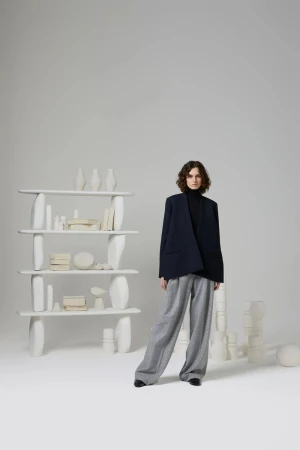 A woman in a navy blazer and grey pants stands in a photo studio setting with an open shelf and various white decorative objects.