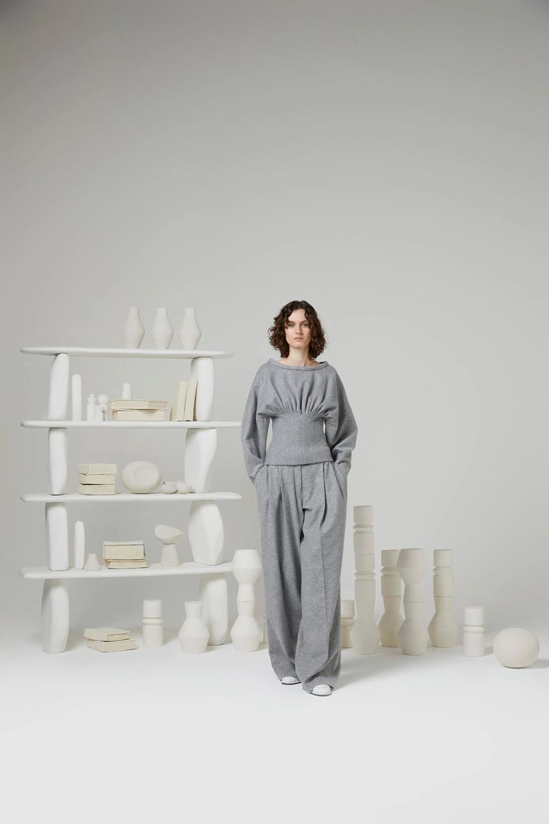 A woman in a grey outfit stands in front of a white shelf filled with vases and books in a bright studio, fashion shot.