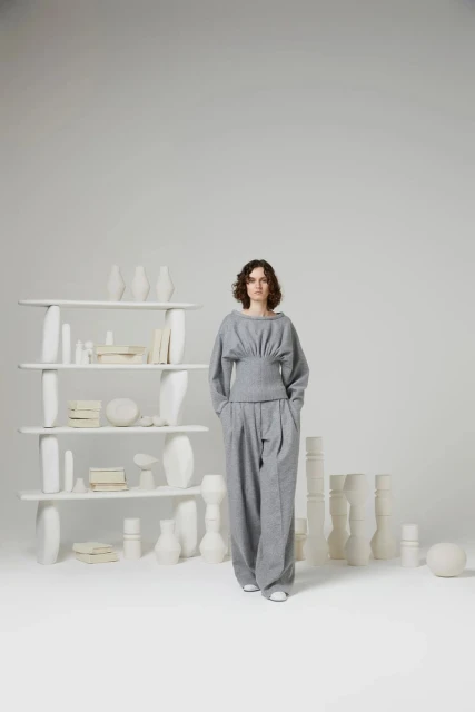 A woman in a grey outfit stands in front of a white shelf filled with vases and books in a bright studio, fashion shot.
