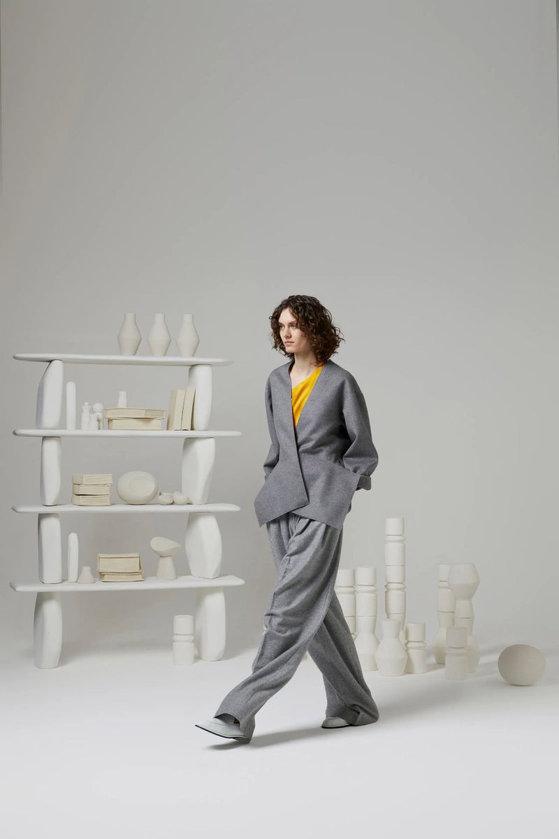 A woman in a grey pant suit walks in a photo studio with white minimalist display shelves and vase-like objects in the background.