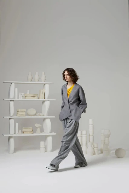 A woman in a grey pant suit walks in a photo studio with white minimalist display shelves and vase-like objects in the background.