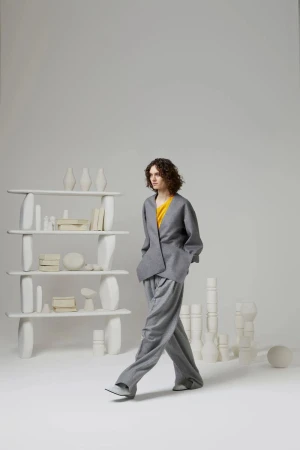 A woman in a grey pant suit walks in a photo studio with white minimalist display shelves and vase-like objects in the background.
