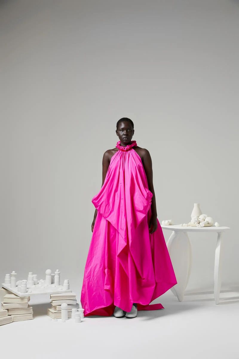A woman poses in a bright pink gown with a high collar in a studio setting, alongside a white table and objects on the floor.