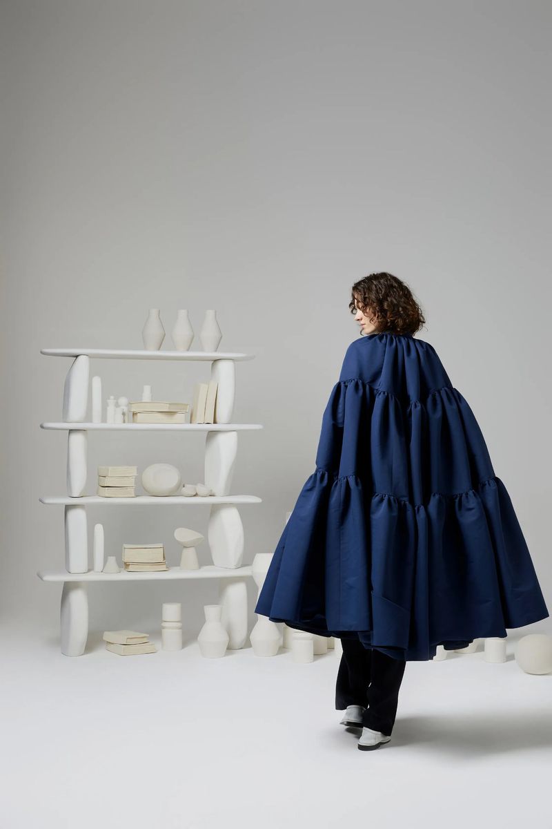 Woman in a voluminous, tiered blue dress walks past a modern white bookshelf filled with white vases and decorative objects in a bright studio.