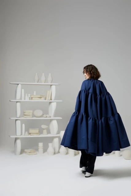 Woman in a voluminous, tiered blue dress walks past a modern white bookshelf filled with white vases and decorative objects in a bright studio.