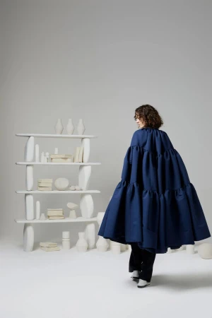 Woman in a voluminous, tiered blue dress walks past a modern white bookshelf filled with white vases and decorative objects in a bright studio.