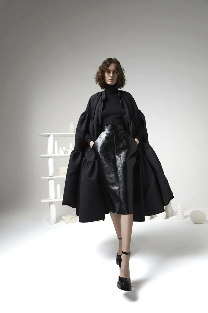 A woman in a black outfit with an oversized coat poses in a bright studio with a minimalist shelf and white geometric objects.