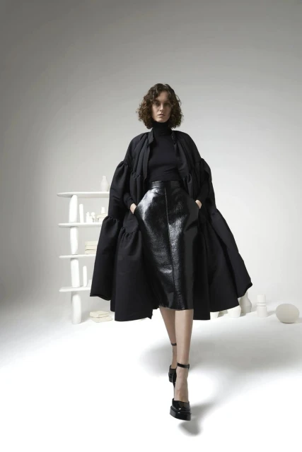 A woman in a black outfit with an oversized coat poses in a bright studio with a minimalist shelf and white geometric objects.