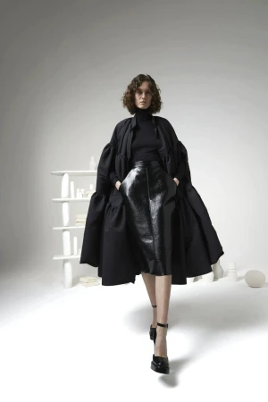 A woman in a black outfit with an oversized coat poses in a bright studio with a minimalist shelf and white geometric objects.