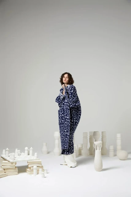 Adult woman in a patterned robe poses in a bright studio with chess and pottery.  Indoor studio lighting, fashion, style, and unique design.
