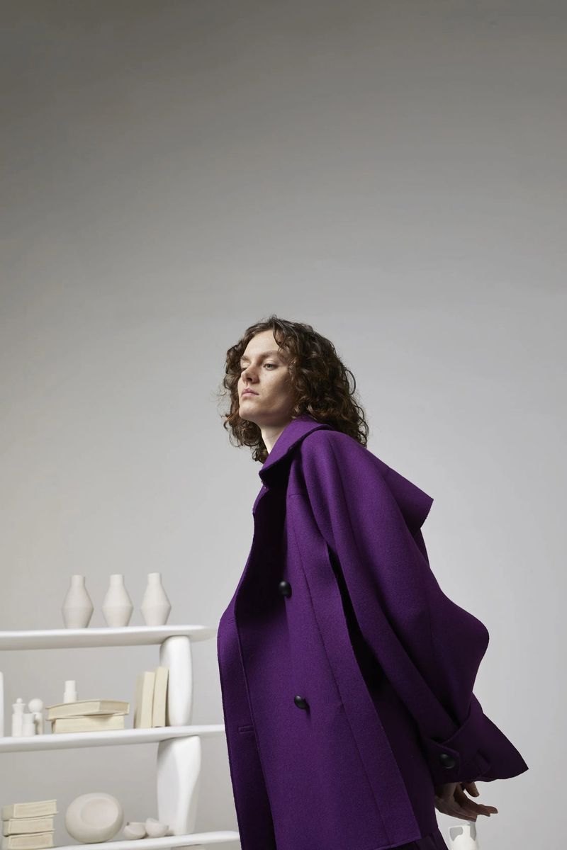 Fashion shoot of a woman with curly brown hair wearing a purple coat, standing next to a white shelving unit filled with white objects.