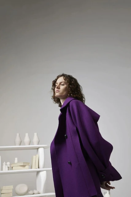 Fashion shoot of a woman with curly brown hair wearing a purple coat, standing next to a white shelving unit filled with white objects.