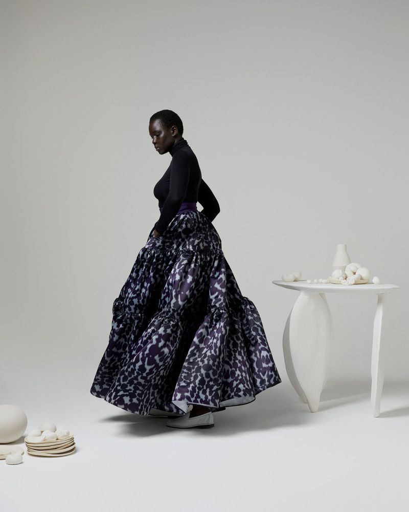 An African woman in a black turtleneck poses in a studio with a large, tiered, patterned skirt, standing near a white table with vases.