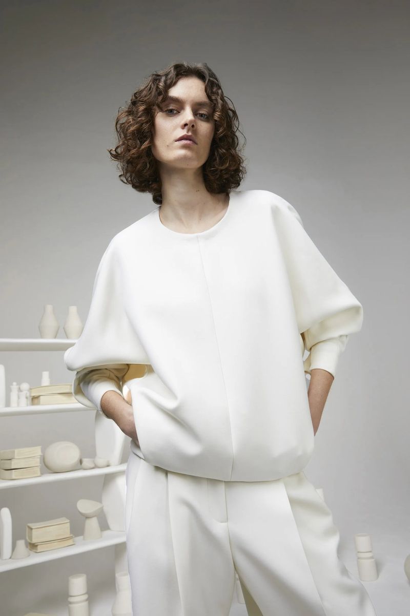 A woman poses in a white oversized sweater and wide-leg trousers in a neutral studio, highlighted by soft, diffused lighting.